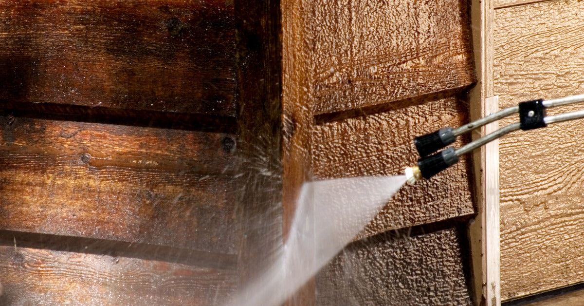 A pressure washer spraying water across dark wooden siding beside lighter textured exterior wall panels.