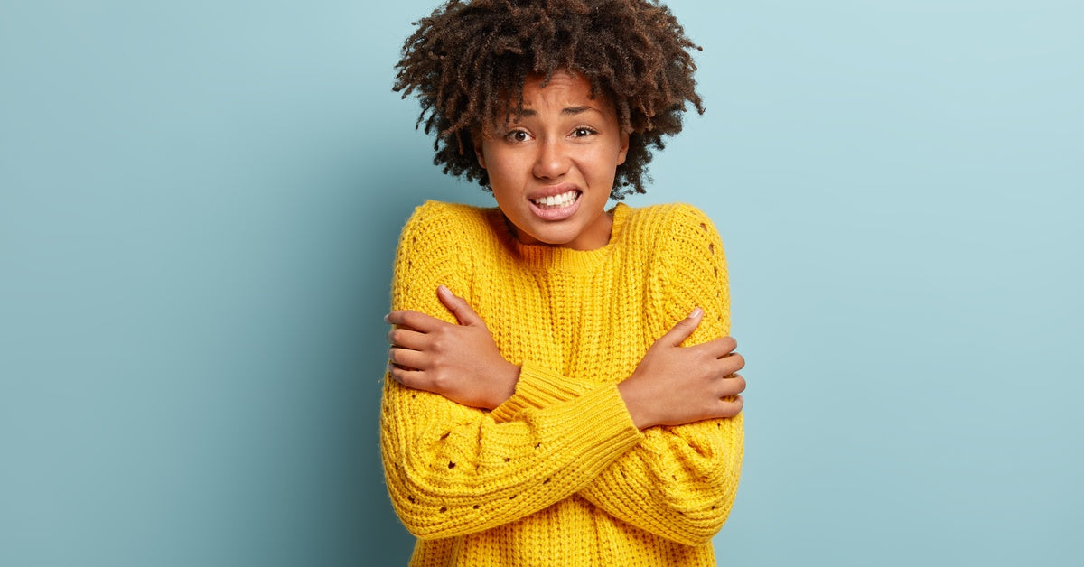 A woman wearing a yellow knitted sweater standing against a blue backdrop and shivering with her arms around her chest.