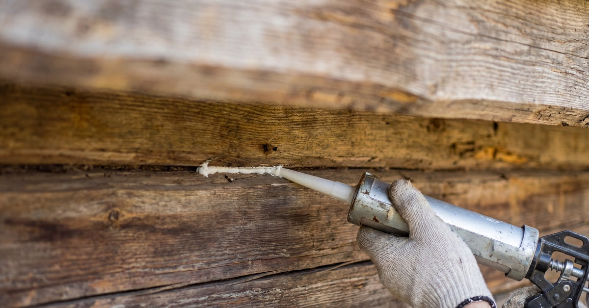 A gloved hand using a caulking gun to apply white sealant between weathered horizontal wooden logs closely.