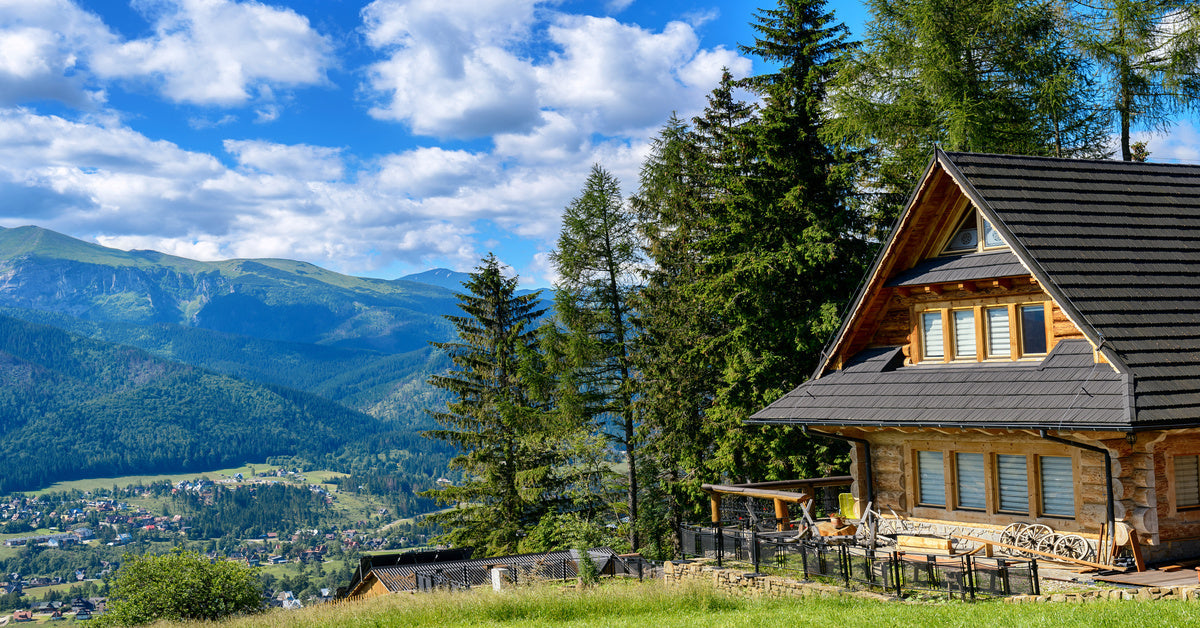 A log cabin perched on a wooded cliff, offering views of a town below and mountains in the distance.