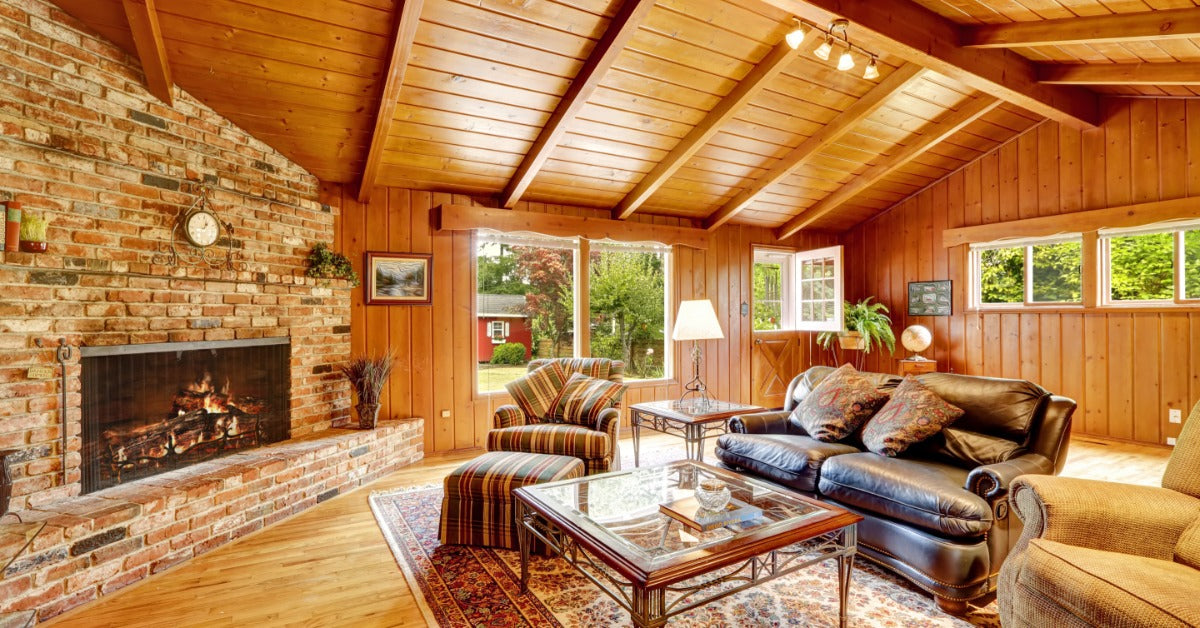 A wood-paneled living room with a brick fireplace, leather sofa, armchairs, rug, and large bright windows.