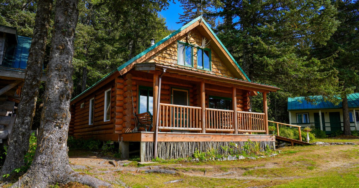 A log cabin with a covered porch standing among tall evergreen trees and grass under a clear blue sky.